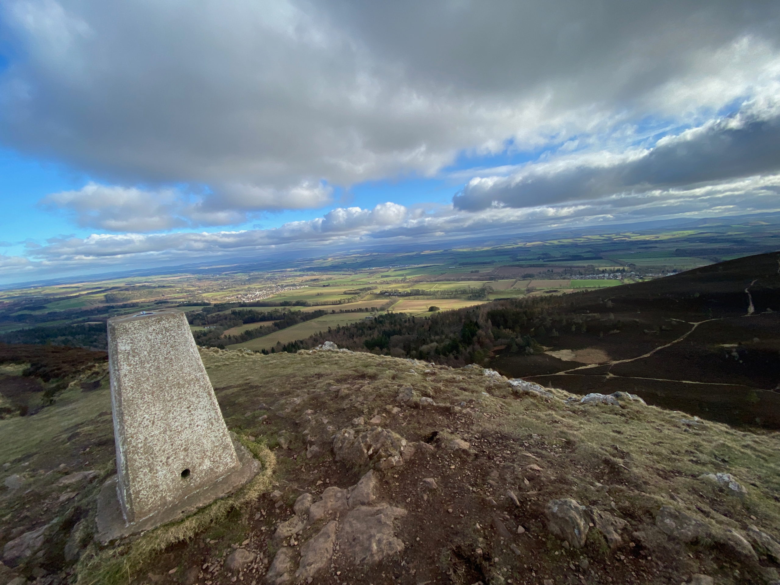 A trig point at the top of a hill with an open view stretching out below